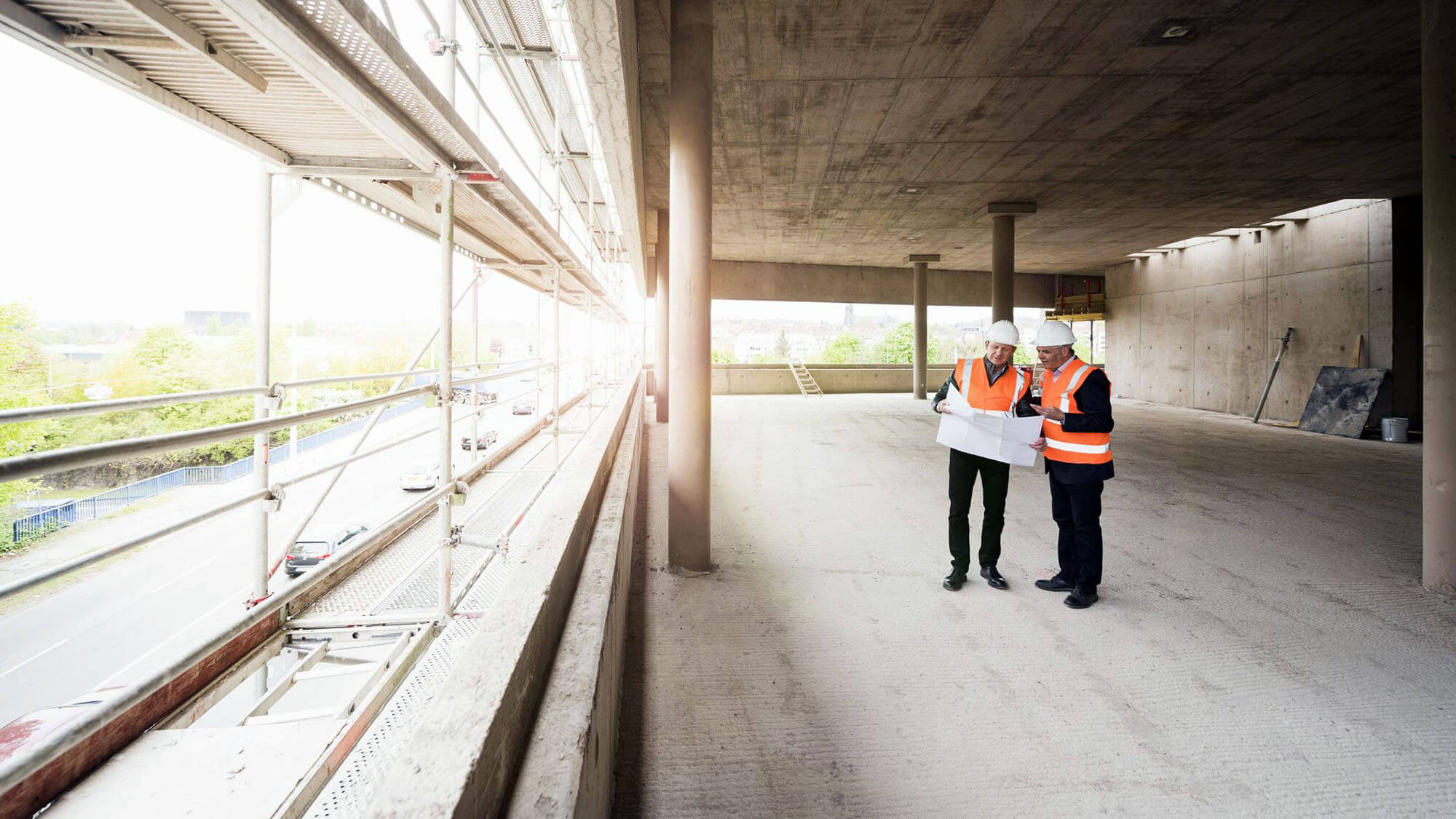 Two men with plan wearing safety vests talking in building under construction 